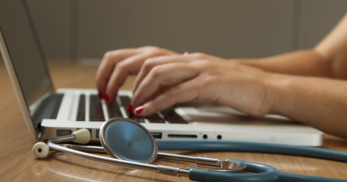 Nursing professional typing on a laptop with a stethoscope beside them on a desk