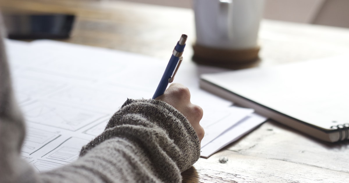 Student writes notes at a desk with a coffee mug and notebook for exam preparation